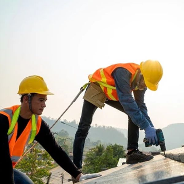 Two construction workers wearing hard hats and safety harnesses installing rooftop safety equipment on a commercial roof.