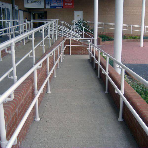 An ADA-compliant metal handrail system installed alongside an outdoor concrete access ramp and brick building.