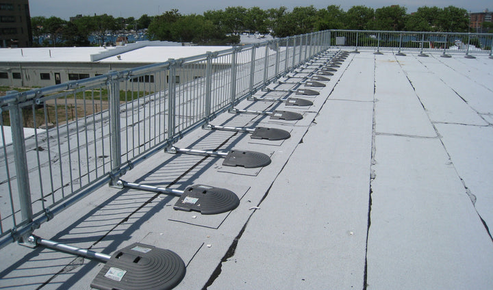 Line of metal non-penetrating guardrail barriers with weighted bases extending along the perimeter of a large flat rooftop.