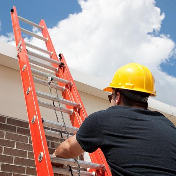 Man wearing a yellow hard hat looking up to a roof holding an orange extension ladder.