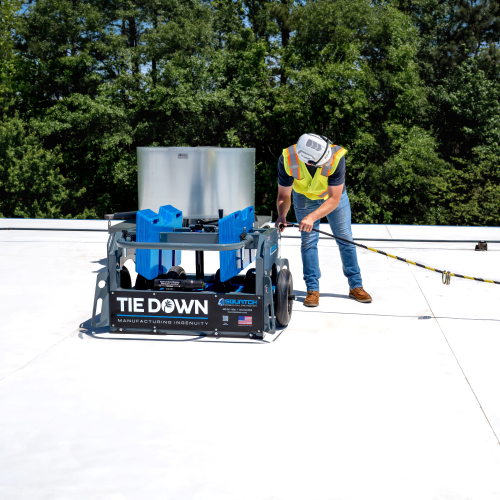 Worker wearing a hard hat and safety harness operating a Tie Down mobile fall protection system cart on a flat commercial roof.