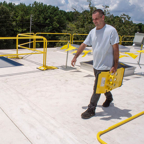 Worker carrying a yellow railing base across a roof.