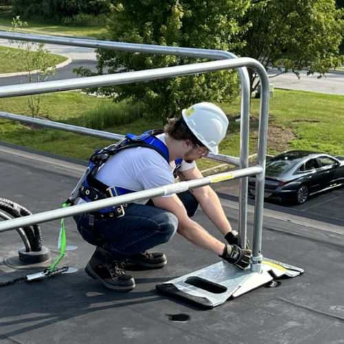 Worker wearing a hard hat and safety harness assembling a portable safety guardrail system on a flat roof.