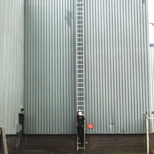 Worker in hard hat and harness climbing a tall, exterior fixed ladder
