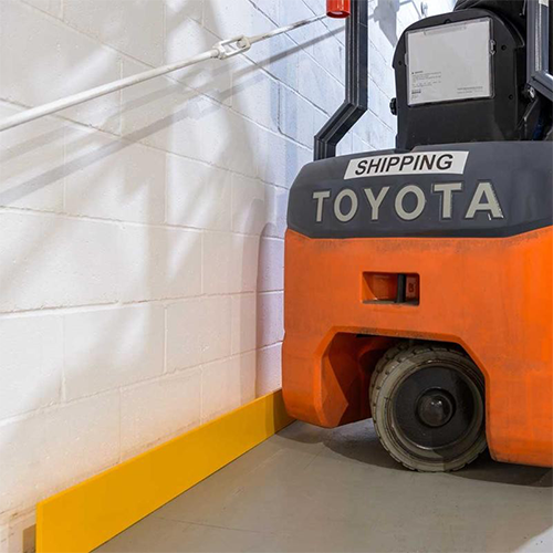 An orange and gray Toyota forklift parked in a warehouse corner next to a white cinderblock wall and a yellow floor bumper.