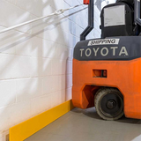 An orange and gray Toyota forklift parked in a warehouse corner next to a white cinderblock wall and a yellow floor bumper.