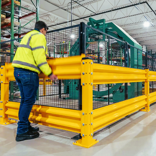 A worker in a high-visibility yellow safety jacket and jeans stands beside a large yellow industrial safety fence with black mesh in a warehouse setting.