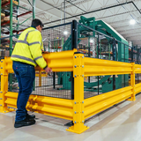 A worker in a high-visibility yellow safety jacket and jeans stands beside a large yellow industrial safety fence with black mesh in a warehouse setting.