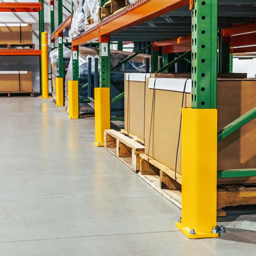 Yellow warehouse pallet bumpers installed on the base of green metal shelving columns in a storage facility, with cardboard boxes on wooden pallets.