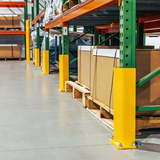 Yellow warehouse pallet bumpers installed on the base of green metal shelving columns in a storage facility, with cardboard boxes on wooden pallets.