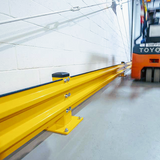 A bright yellow industrial guardrail or channel rail mounted along a warehouse floor against a white brick wall, with part of an orange Toyota forklift visible in the background.