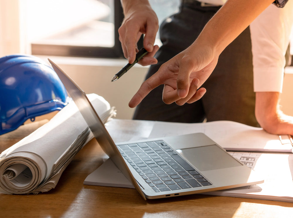 Person pointing at a laptop with construction plans and a hard hat on a desk