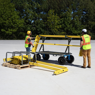 Two workers in safety vests and hard hats operating a large yellow piece of equipment on a clear day with trees in the background.